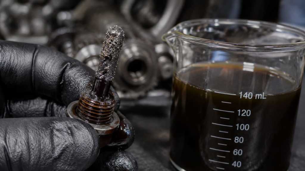 A close-up view of a mechanic's gloved hand holding a Tesla magnetic drain plug coated in fine, shimmering silver metallic sediment, beside a clear beaker of dark, contaminated transmission fluid.